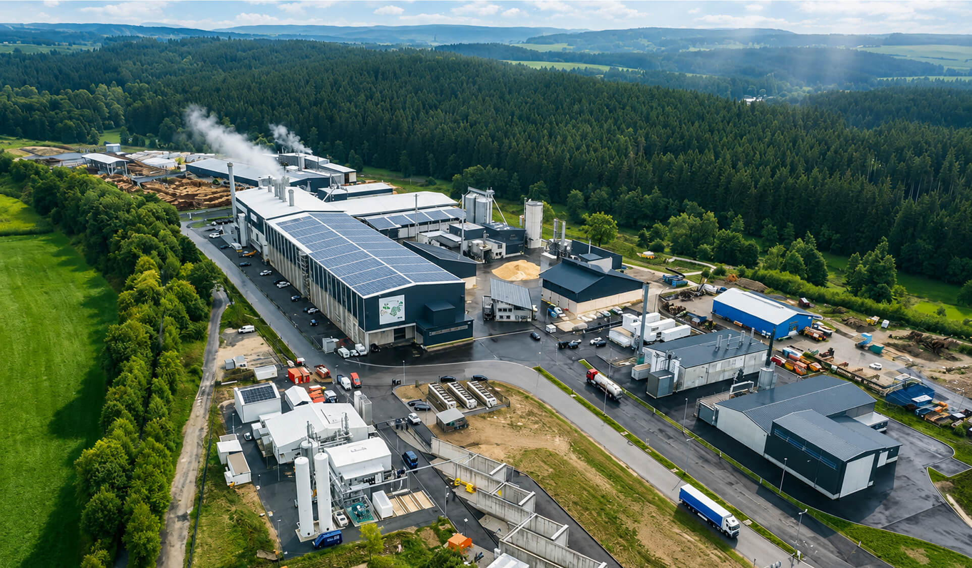 Luftaufnahme des Energieparks Wunsiedel mit mehreren Industriegebäuden, Solarpaneelen auf einem Dach, LKWs und umgeben von Wald und Feldern.