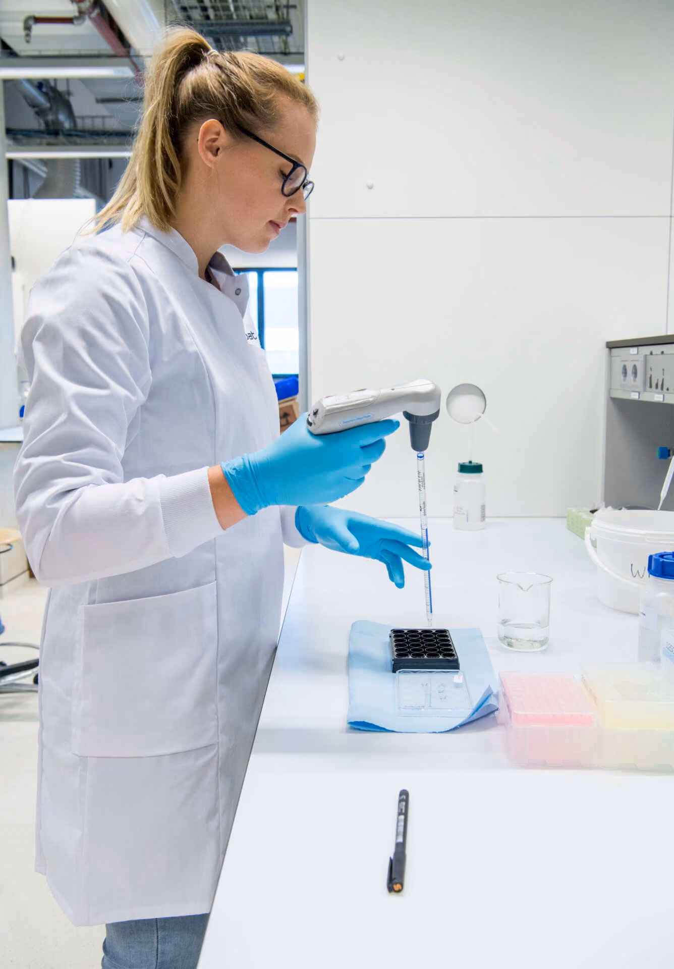 Scientist wearing white lab coat, blue gloves, and glasses using a pipette to transfer liquid into a multi-well plate in a laboratory setting.