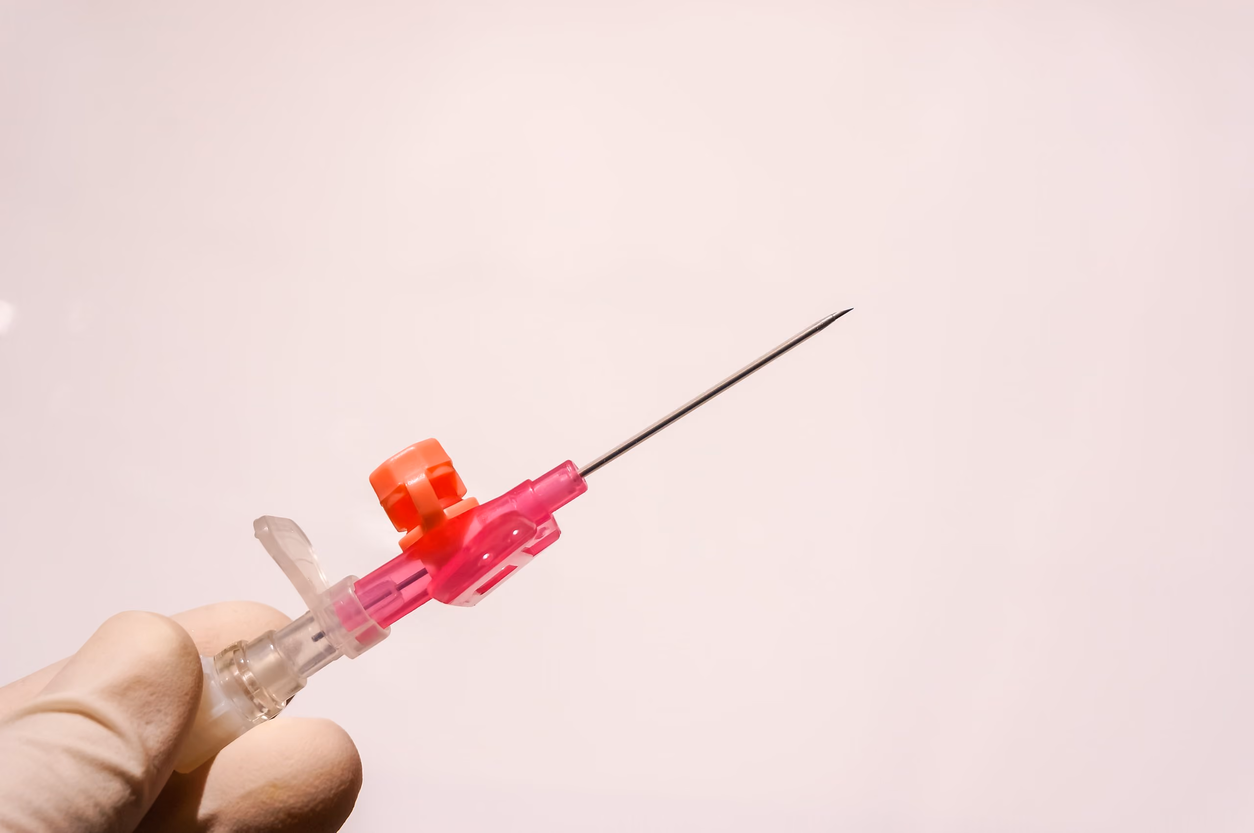 Gloved hand holding a medical needle attached to a pink and clear plastic syringe hub against a plain light background.