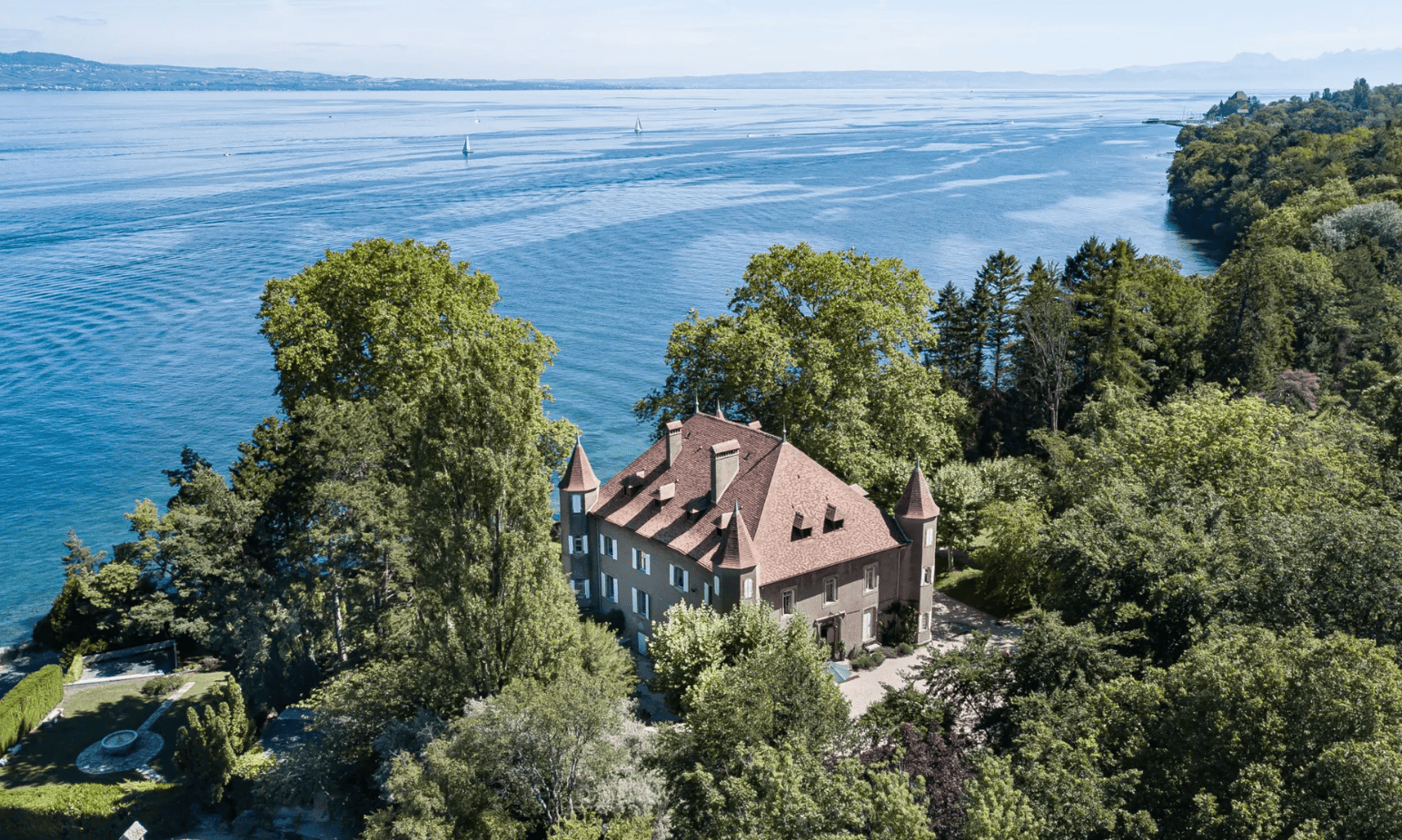 Manoir avec toits en pente entouré d’arbres au bord d’un grand lac bleu avec des voiliers.