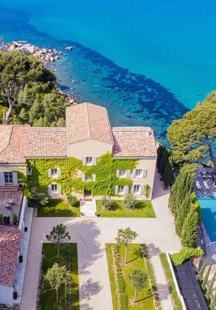 Vue aérienne d'une maison bordée de plantes grimpantes près d'une mer turquoise et entourée d'arbres verts.