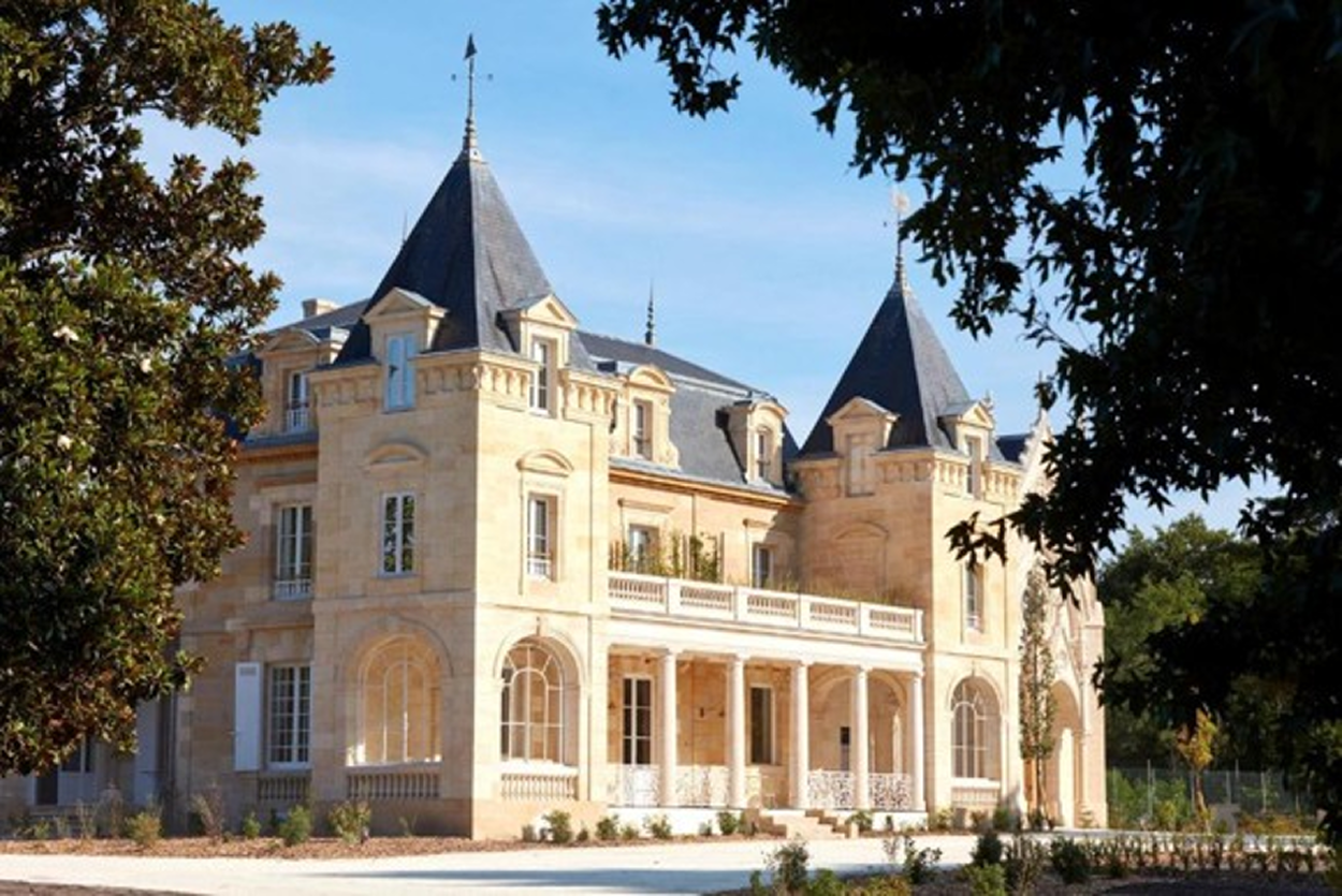 Un château en pierre beige avec deux tours et un porche à colonnes sous un ciel bleu clair.