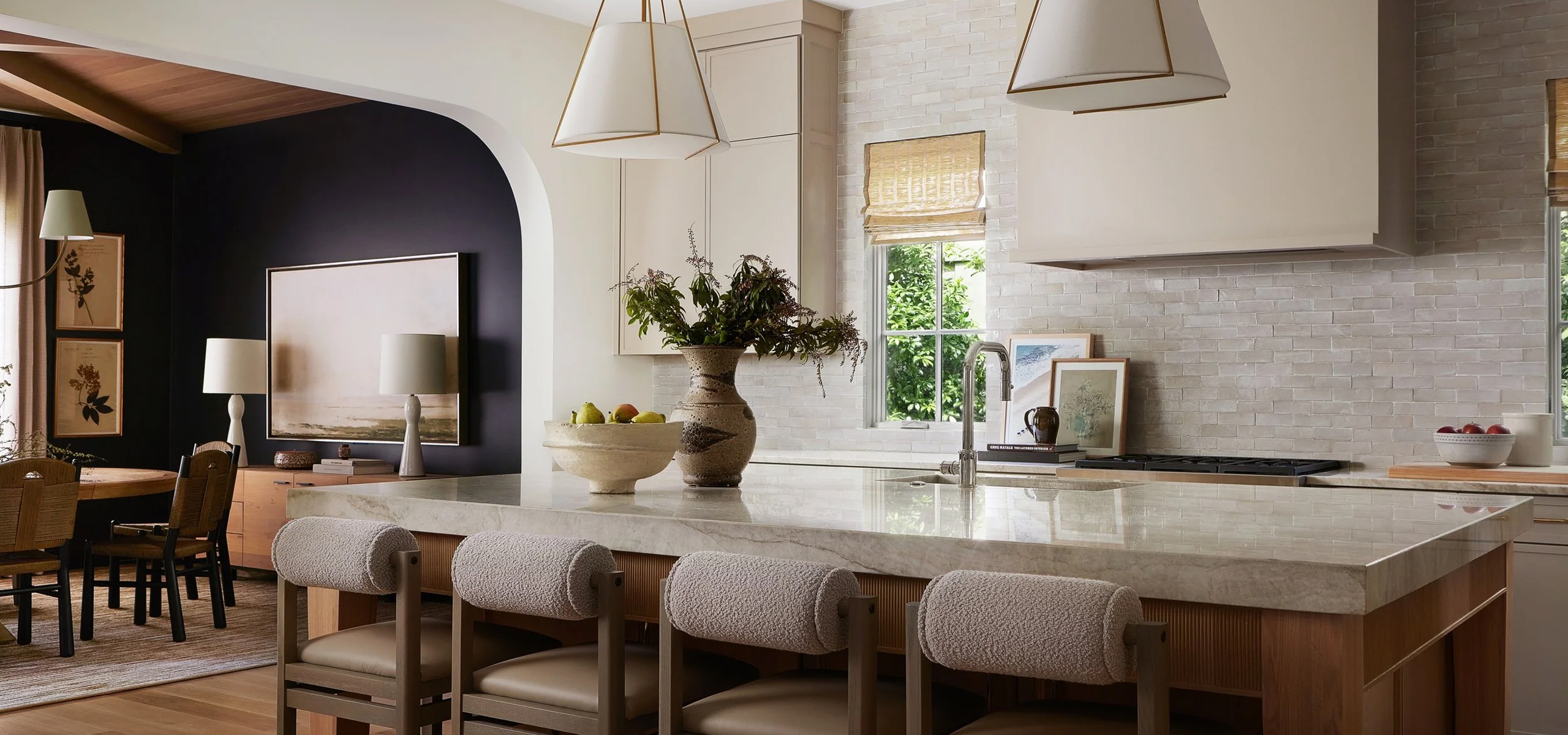 Modern kitchen with marble island, four cushioned stools, pendant lights, and fruit bowls on counter.