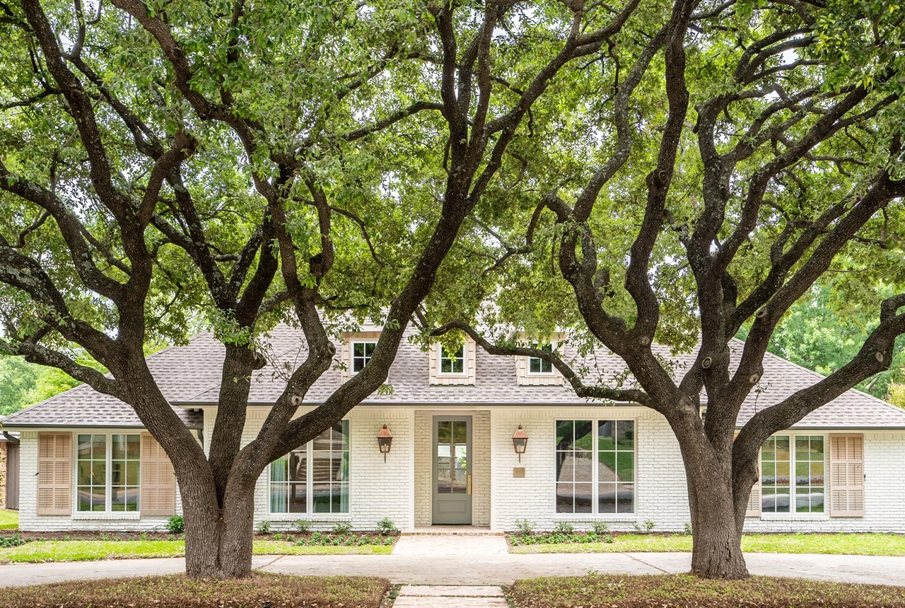 White brick house with large windows, a central door, and two large leafy trees in front.