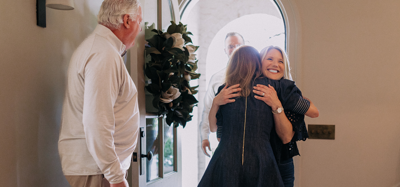 Two women hugging happily near an open door while two men look on in a warmly lit home entryway.