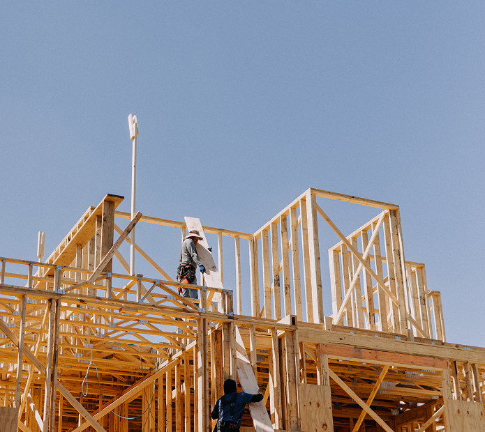 Two construction workers assembling wooden framing of a house under clear blue sky.