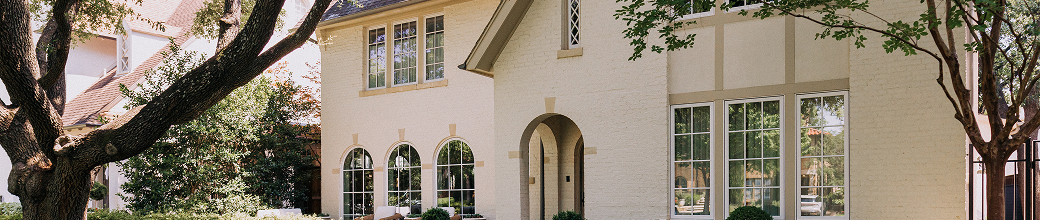 Exterior view of a light-colored house with arched and rectangular windows, surrounded by trees.