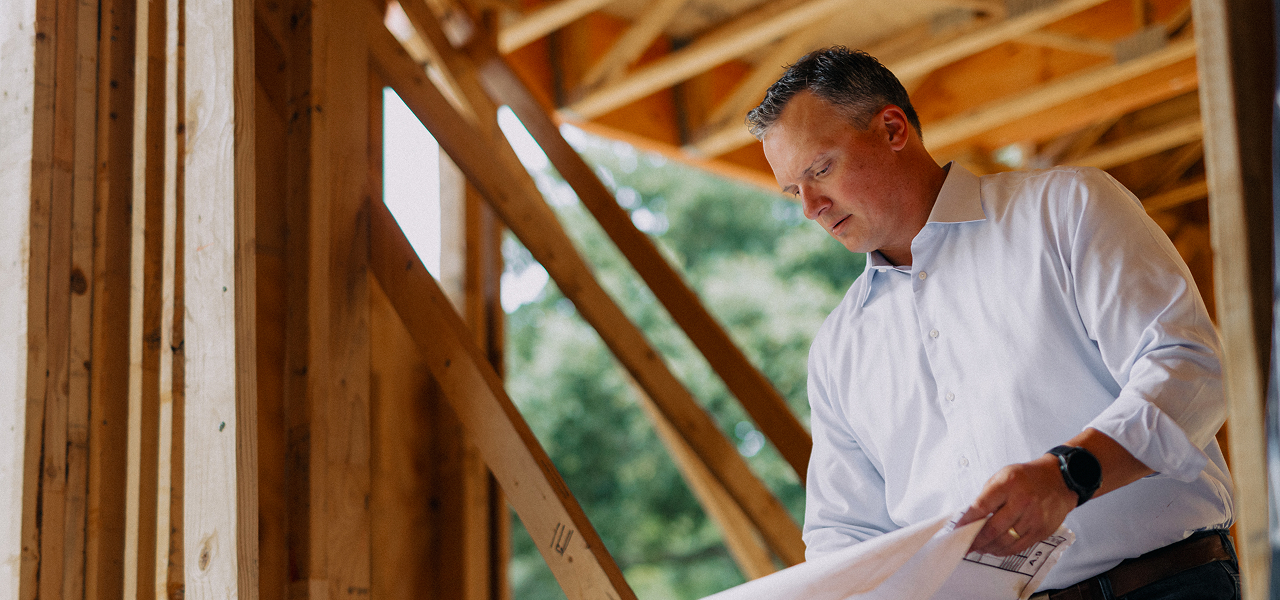 Man in white shirt reviewing construction blueprints inside a wooden building frame.