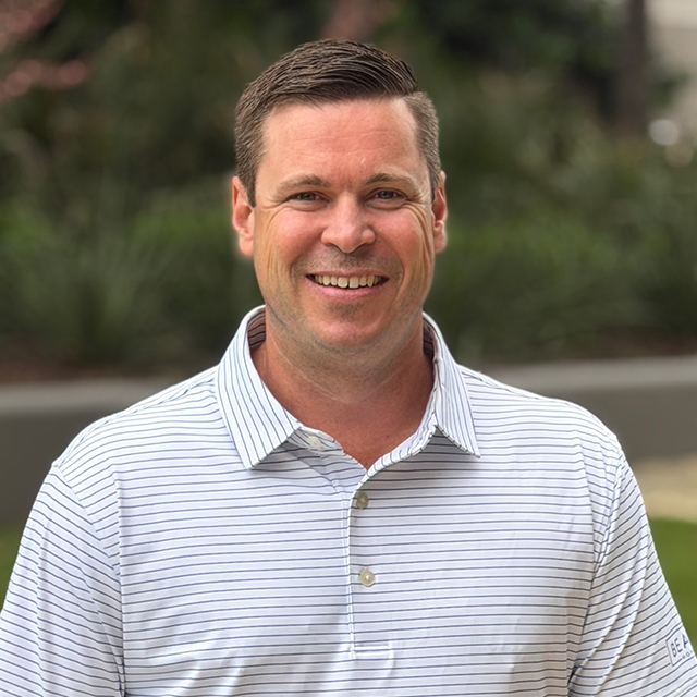 Smiling man with short dark hair wearing a white striped polo shirt outdoors.
