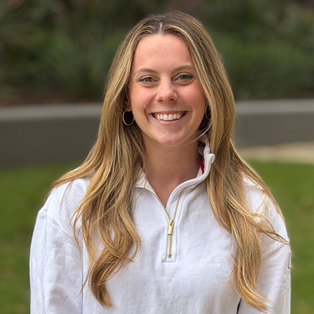 Smiling woman with long blonde hair wearing a white zip-up sweatshirt outdoors with greenery in background.