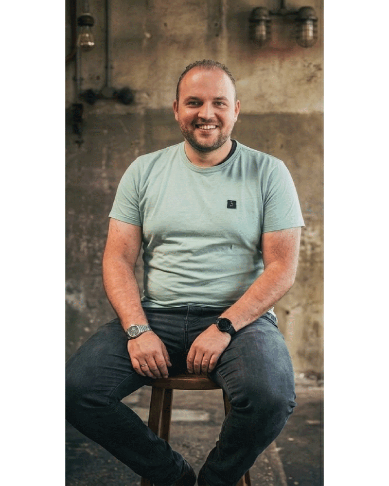 Man met korte haar en baard, zittend op een kruk, glimlachend in een industriële achtergrond.