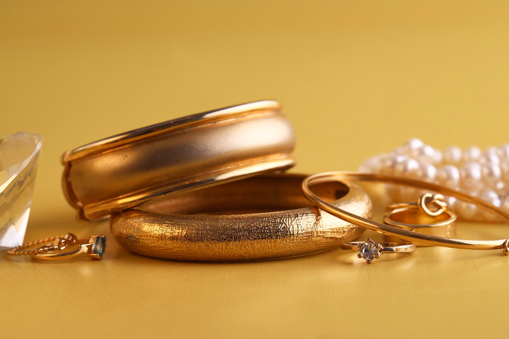 Jóias de ouro com anéis, pulseiras, e colar de pérolas sobre fundo amarelo.
