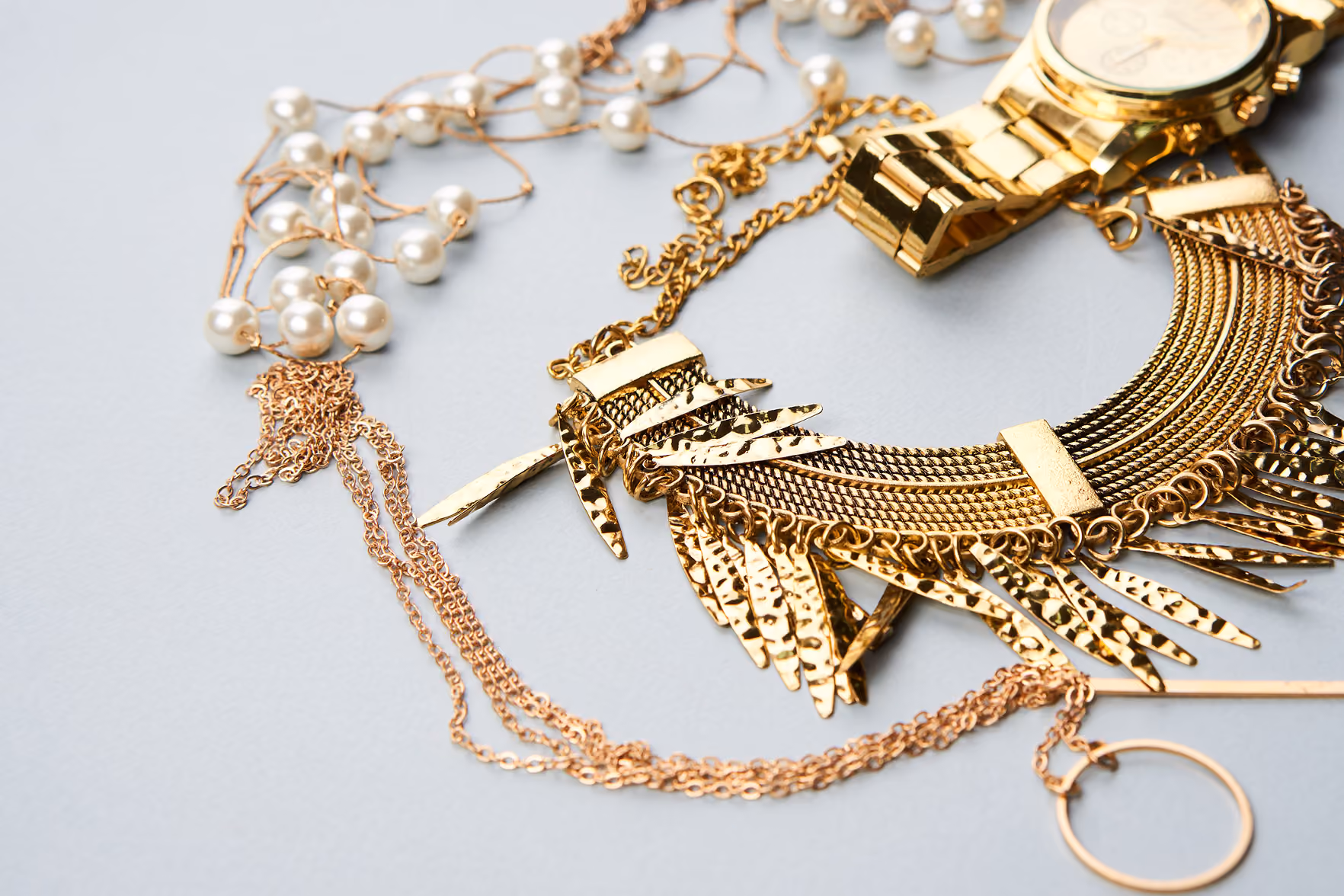Colar dourado decorado com várias barras de metal, pulseira e corrente com pérolas sobre fundo cinza.