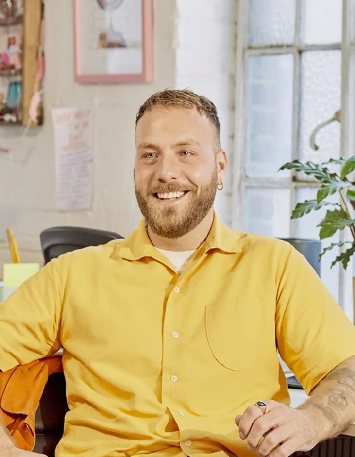 Smiling man with beard and earring wearing a yellow shirt sitting in a bright room with a window.