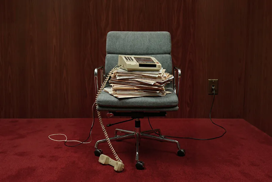 Vintage office chair with a pile of folders and a beige corded telephone, handset off the hook on red carpet.