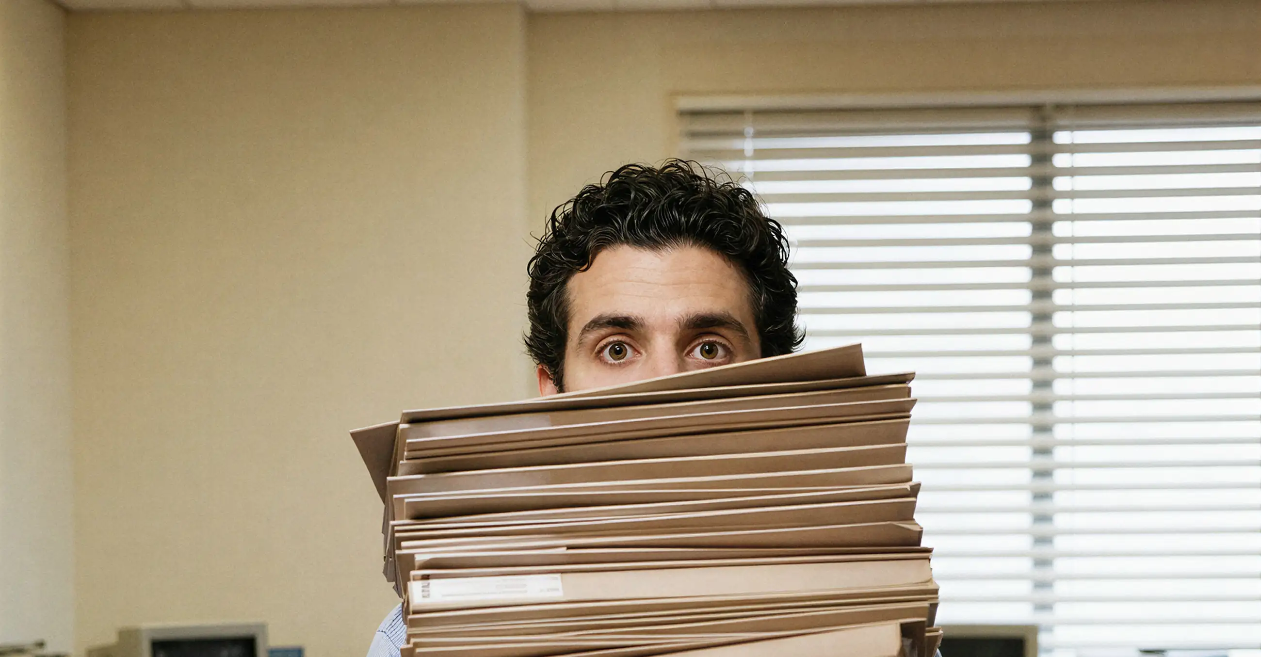 Man in office holding a large stack of file folders, hiding part of his face behind them.