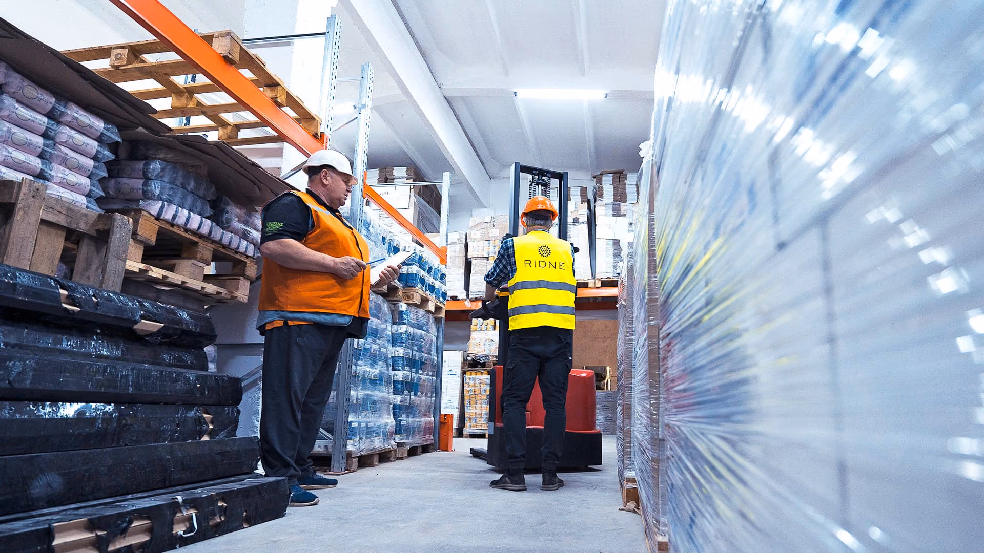 Two warehouse workers wearing safety gear handling pallets in an industrial storage area.
