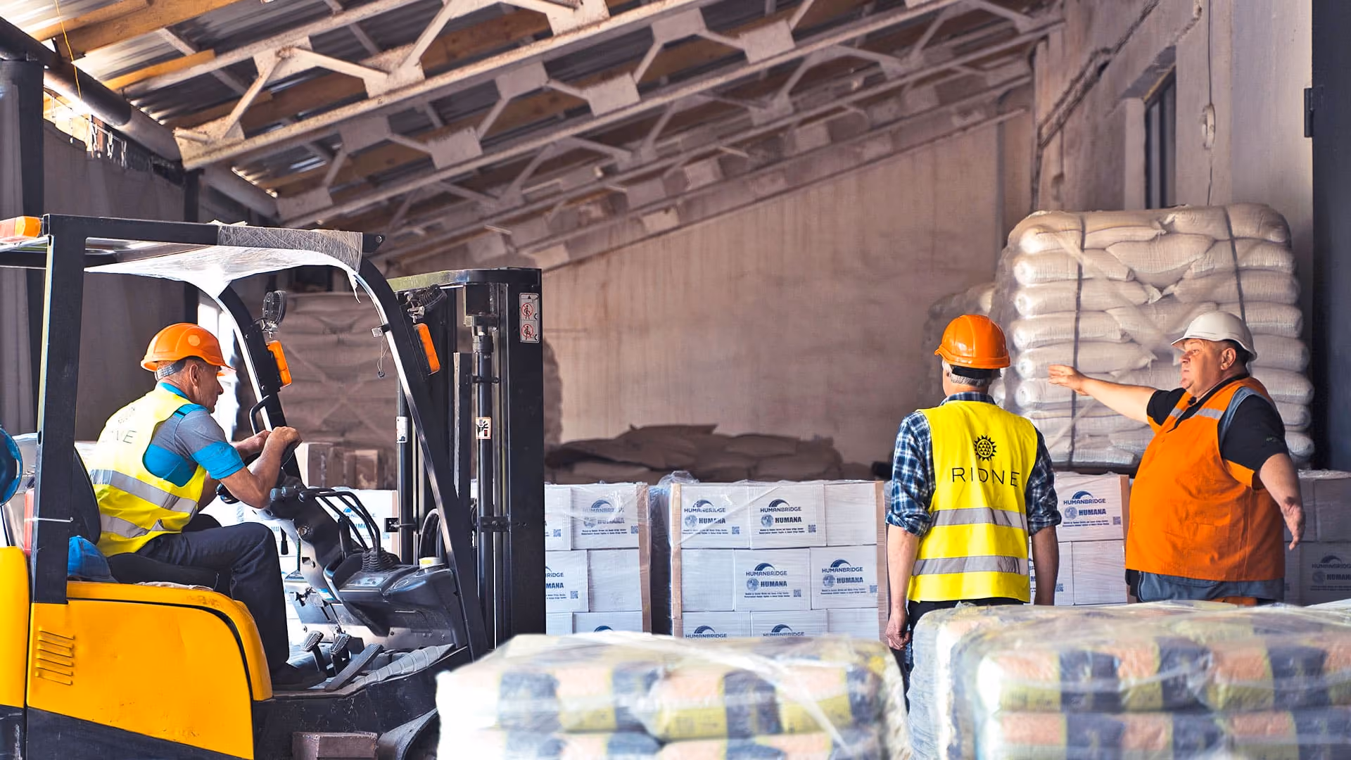 Worker driving forklift in warehouse talks with two colleagues in safety vests and helmets.