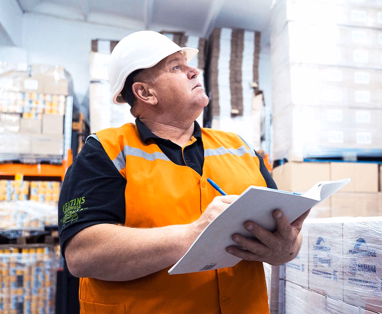 Warehouse worker in orange vest and white hard hat holding a clipboard and pen, inspecting stock.