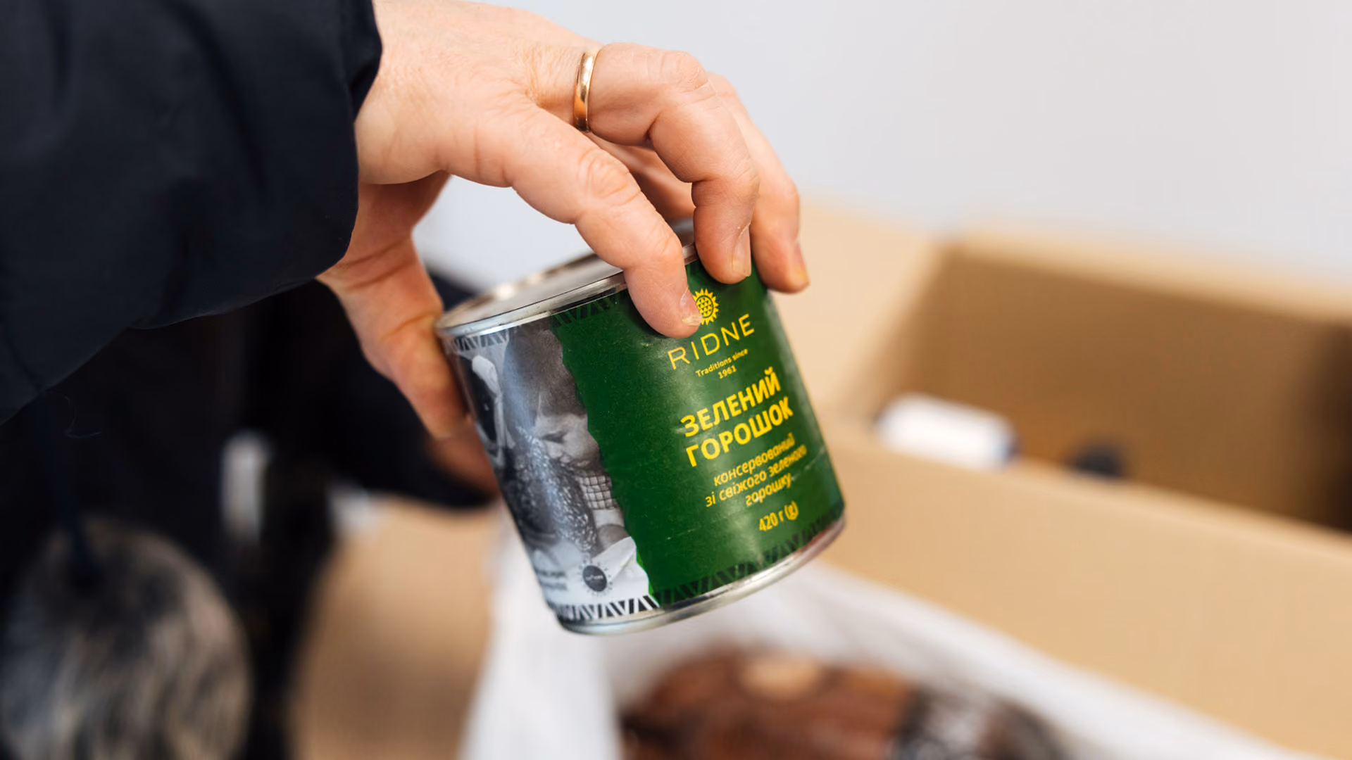 Hand with wedding ring holding a can of Ridne brand green peas over a cardboard box.
