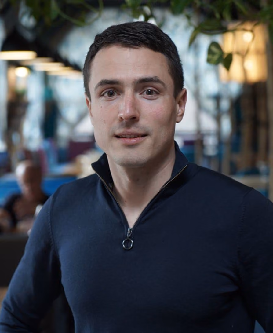Serhiy Kovalchuk with short dark hair wearing a dark zippered shirt indoors with blurred background lighting.