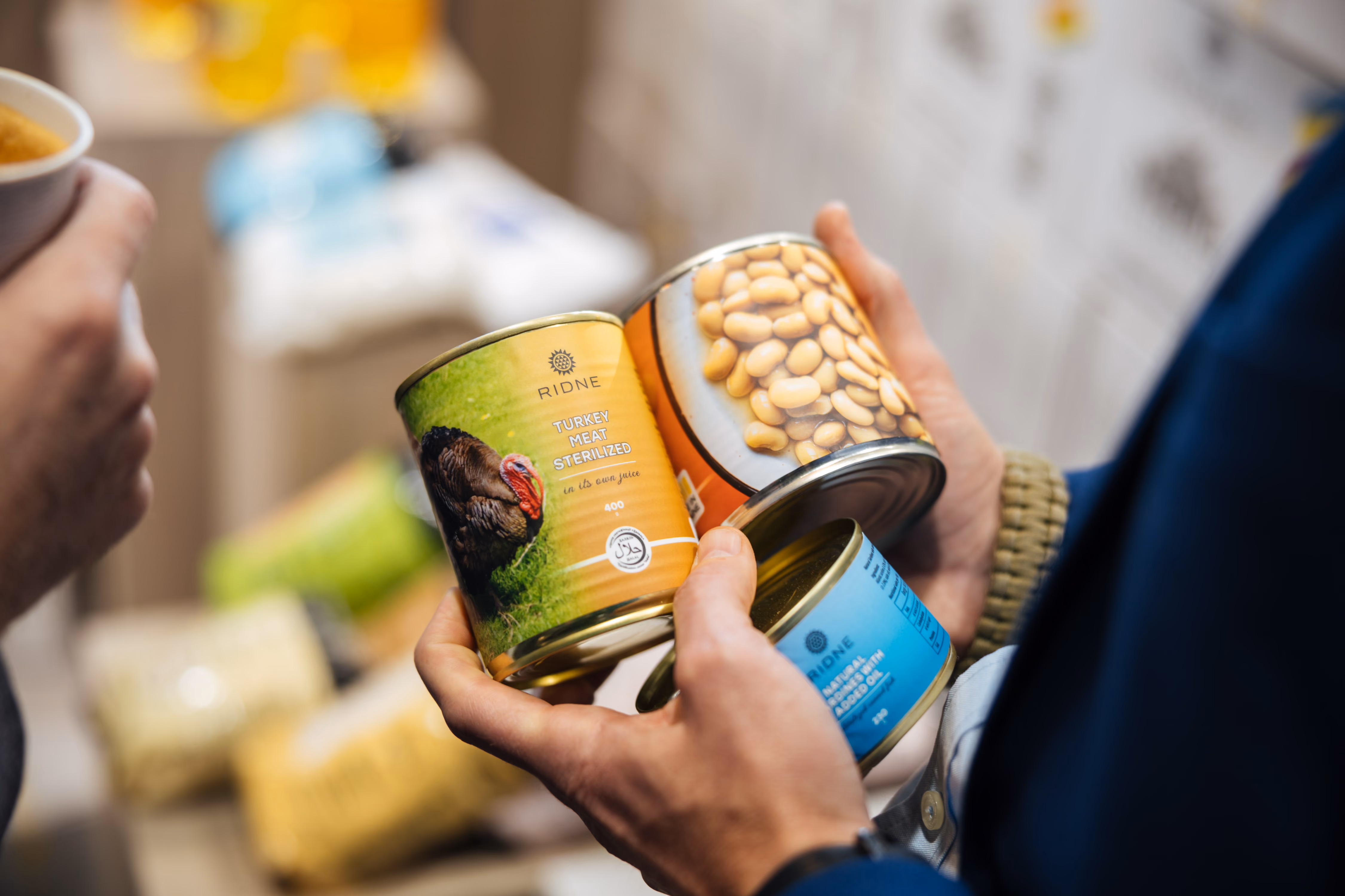 Person holding canned turkey meat, beans, and a blue-labeled can in their hands indoors.