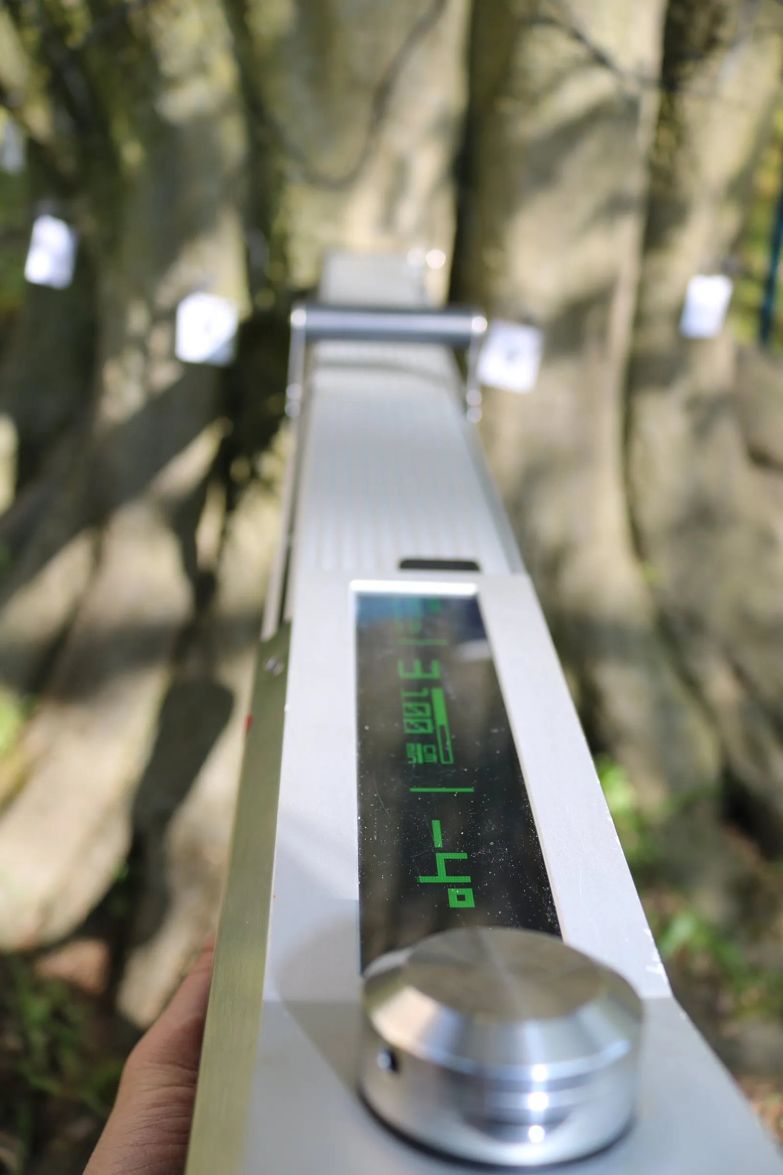 Hand hält ein digitales Messgerät mit grünen Ziffern auf einem Display, das auf natürliche Umgebung mit Baumrinde und Blättern gerichtet ist.