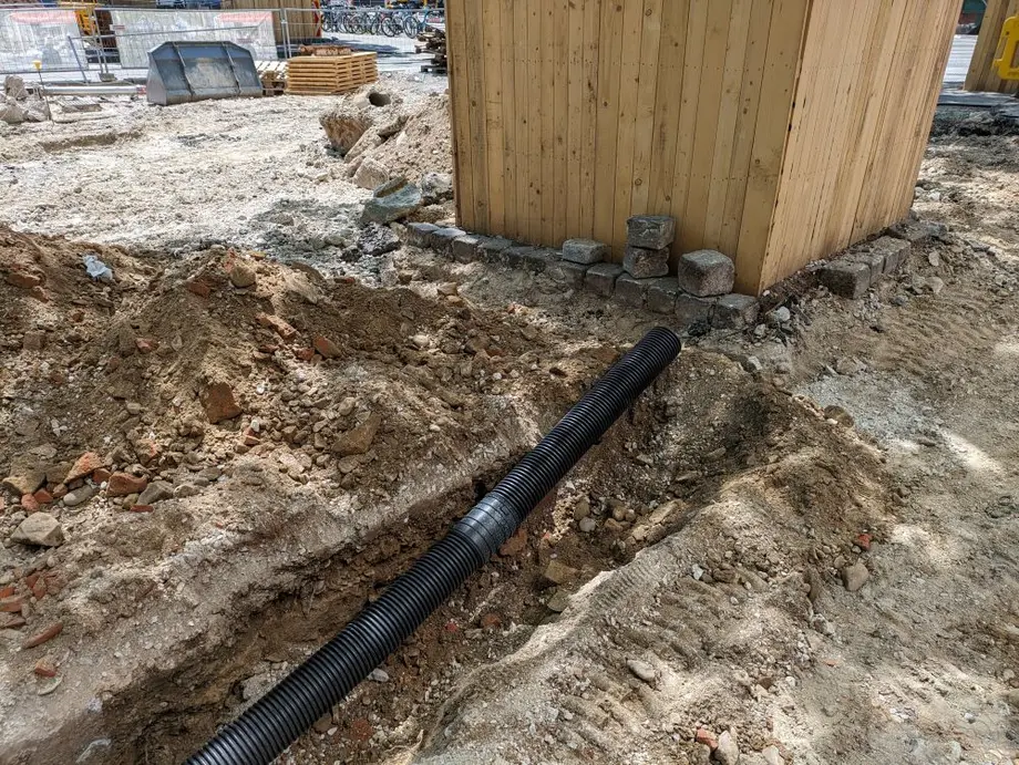 Baustelle mit offener Erde, auf der ein schwarzes Rohr verlegt wird, neben einem Holzcontainer und Baumaterialien im Hintergrund.