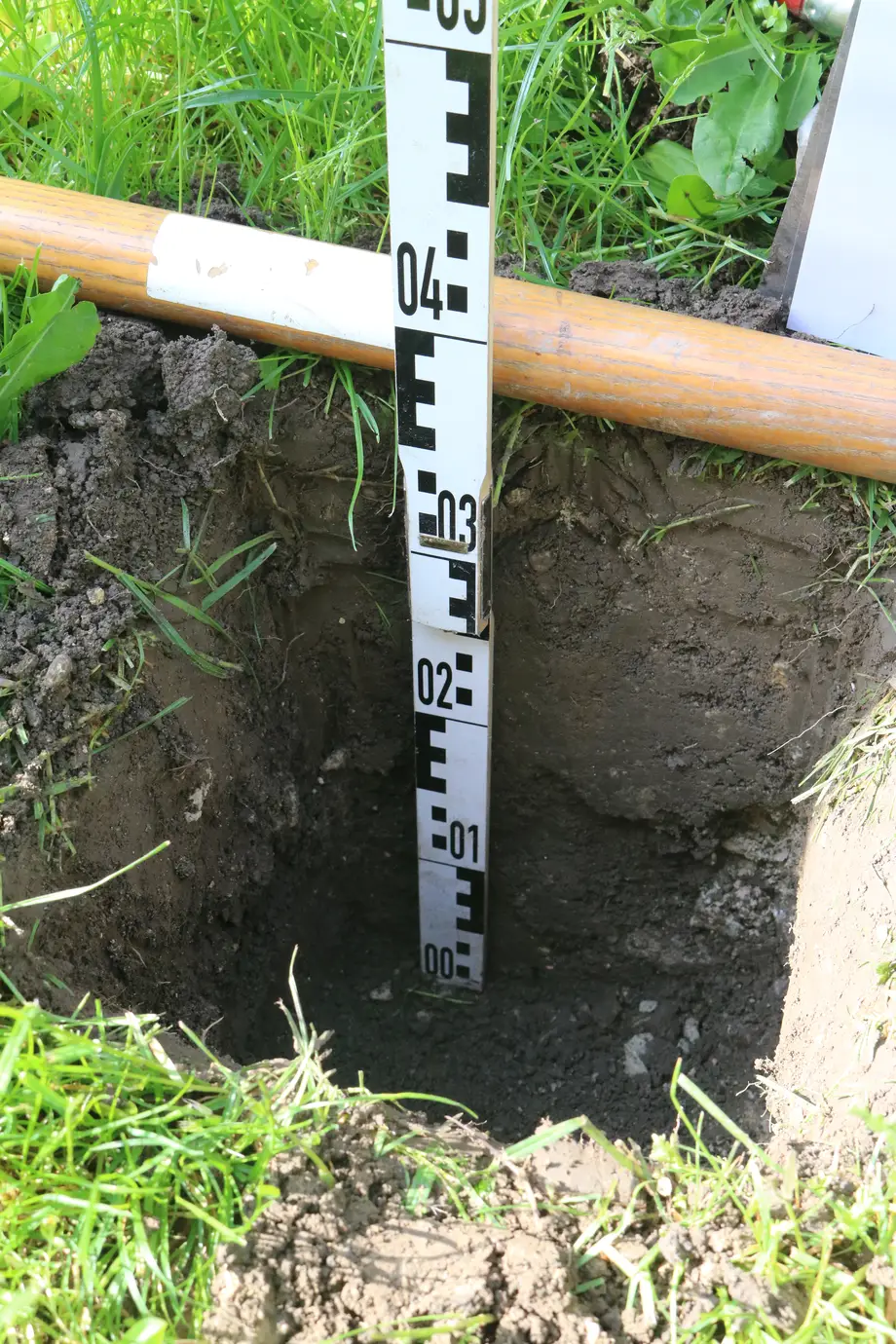 Ein Erdloch mit einem Maßstab, der etwa 0,4 Meter Tiefe anzeigt, umgeben von grünem Gras und einem Holzstab.