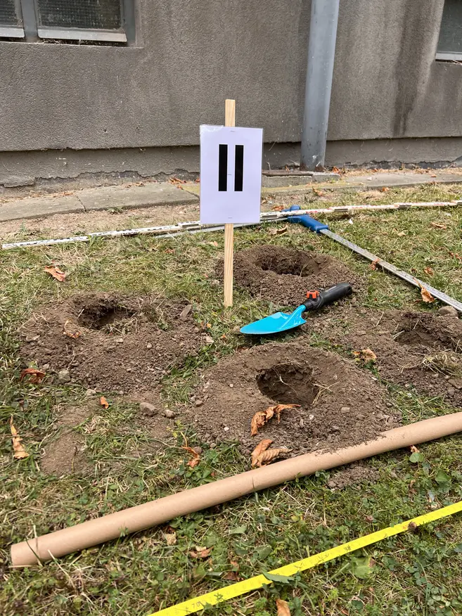 Drei kleine runde Löcher in der Erde auf einem grasbewachsenen Gelände, mit einem blauen Gartenschaufel und einem ausgerollten Maßband daneben. Ein Holzstab mit einem weißen Schild darauf, das zwei schwarze parallele Striche zeigt, steht mittig im Bild. Im Hintergrund ist eine graue Gebäudewand und zwei Fenster sichtbar.