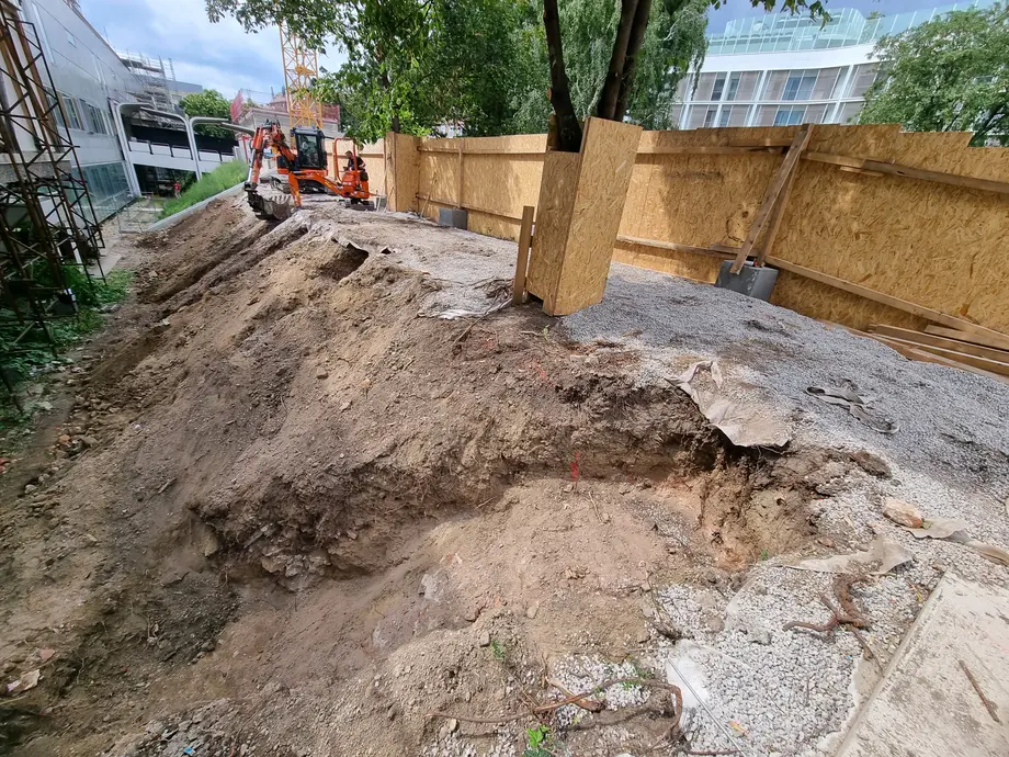 Eine Baustelle mit unebenem, ausgehobenem Erdreich und einem kleinen Bagger, der auf einem schmalen Weg neben einer Holzwand und einem Baum arbeitet.
