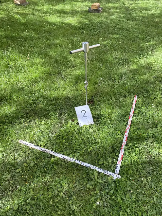 Grüne Wiese mit zwei Maßbändern, die einen rechten Winkel bilden, und einem Metallgerät mit einem T-förmigen Griff, daneben ein Schild mit der Zahl 2.