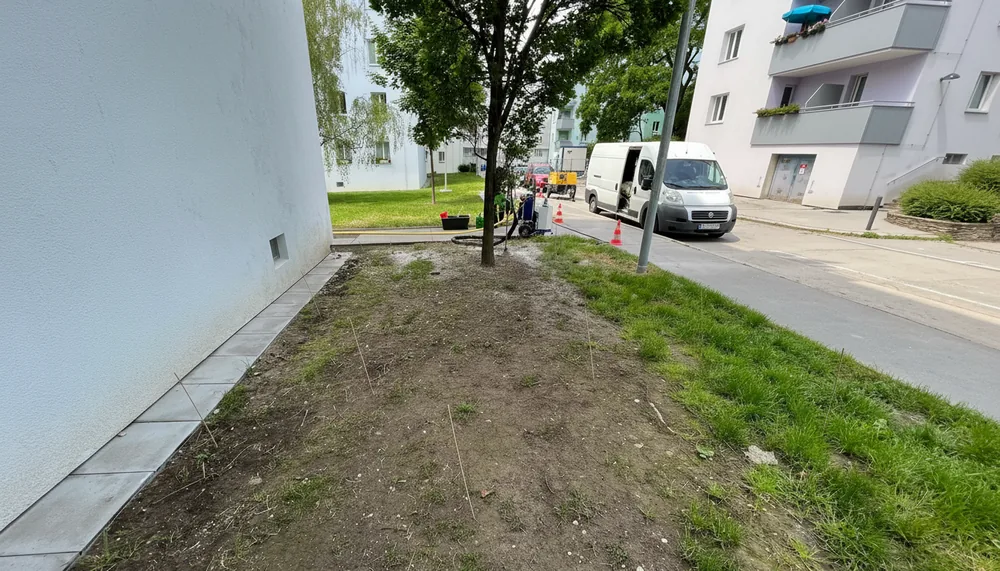 Ein befestigter Gehweg neben einem weißen Gebäude mit kahlem Boden und Grasflächen, rechts eine Straße mit einem weißen Transporter und Baustellenabsperrungen im Hintergrund.