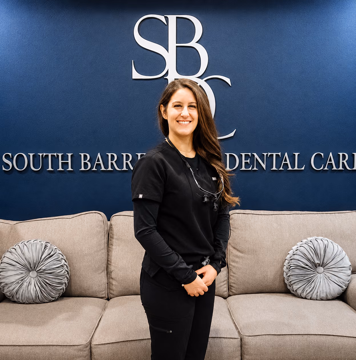 Smiling woman in black dental scrubs stands in front of a sofa with cushions and dental clinic logo.