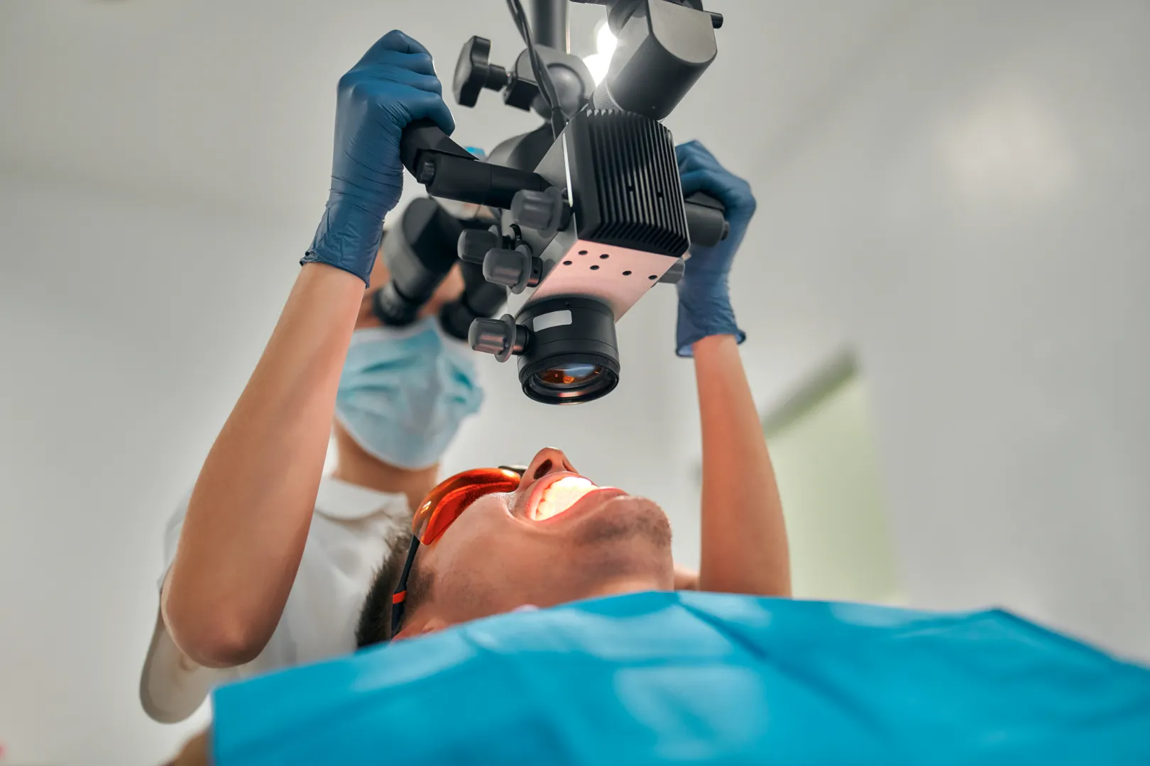 Dentist wearing gloves and mask using a dental microscope to examine a patient’s open mouth.