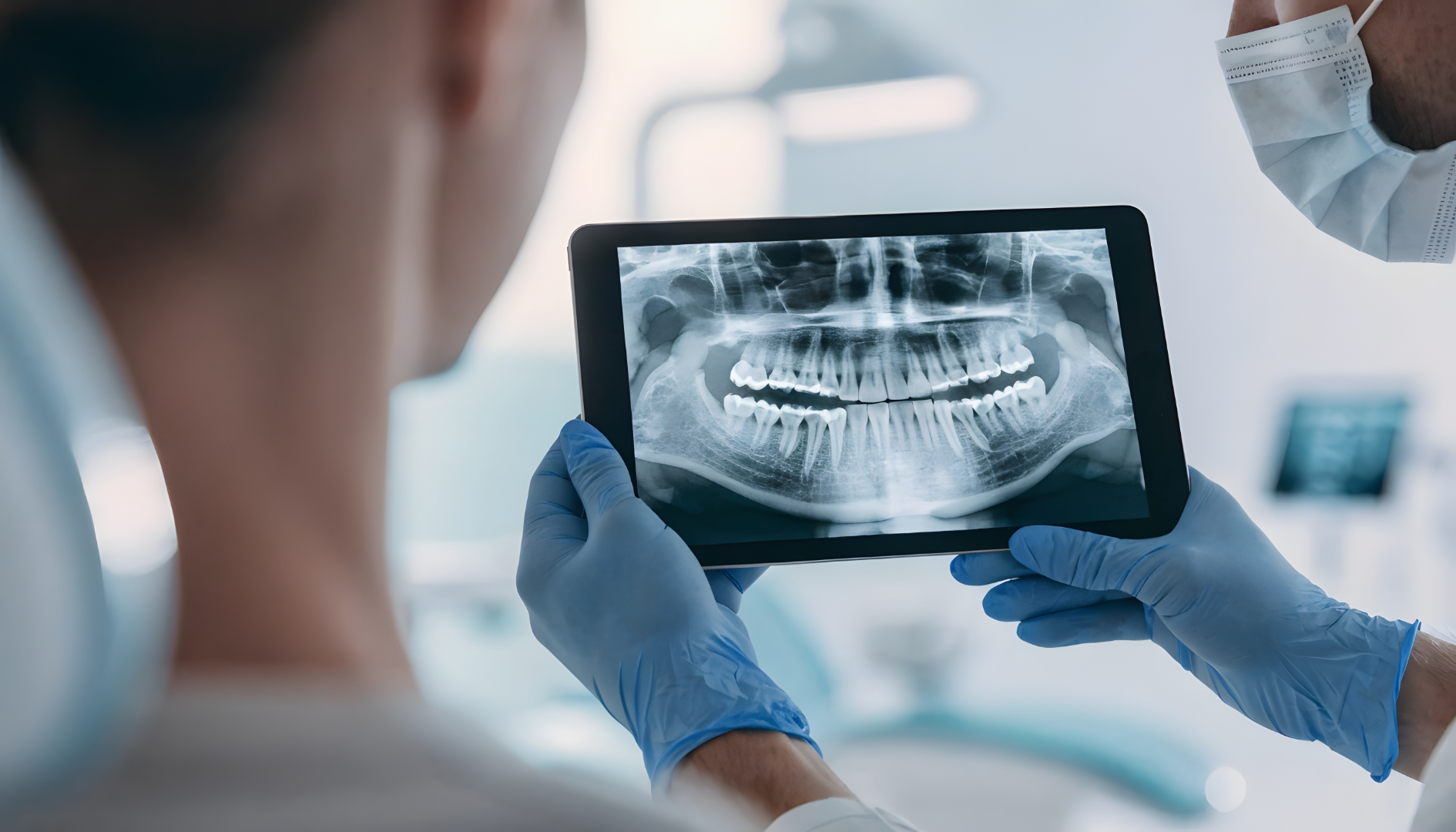 Dentist wearing gloves holding a tablet displaying a dental X-ray of teeth and jawbones.