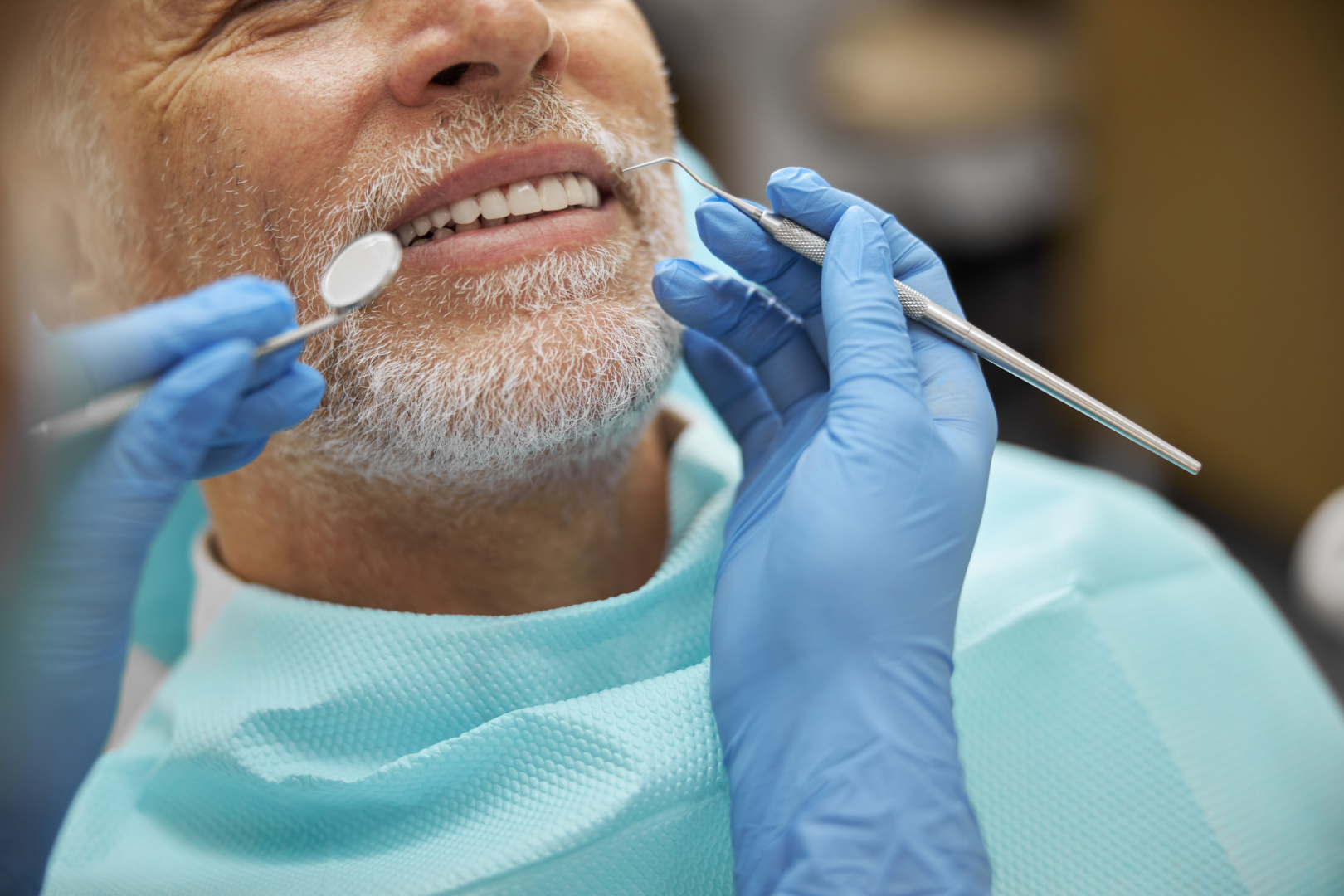 Dentist with blue gloves examining a senior man's teeth using dental mirror and probe.