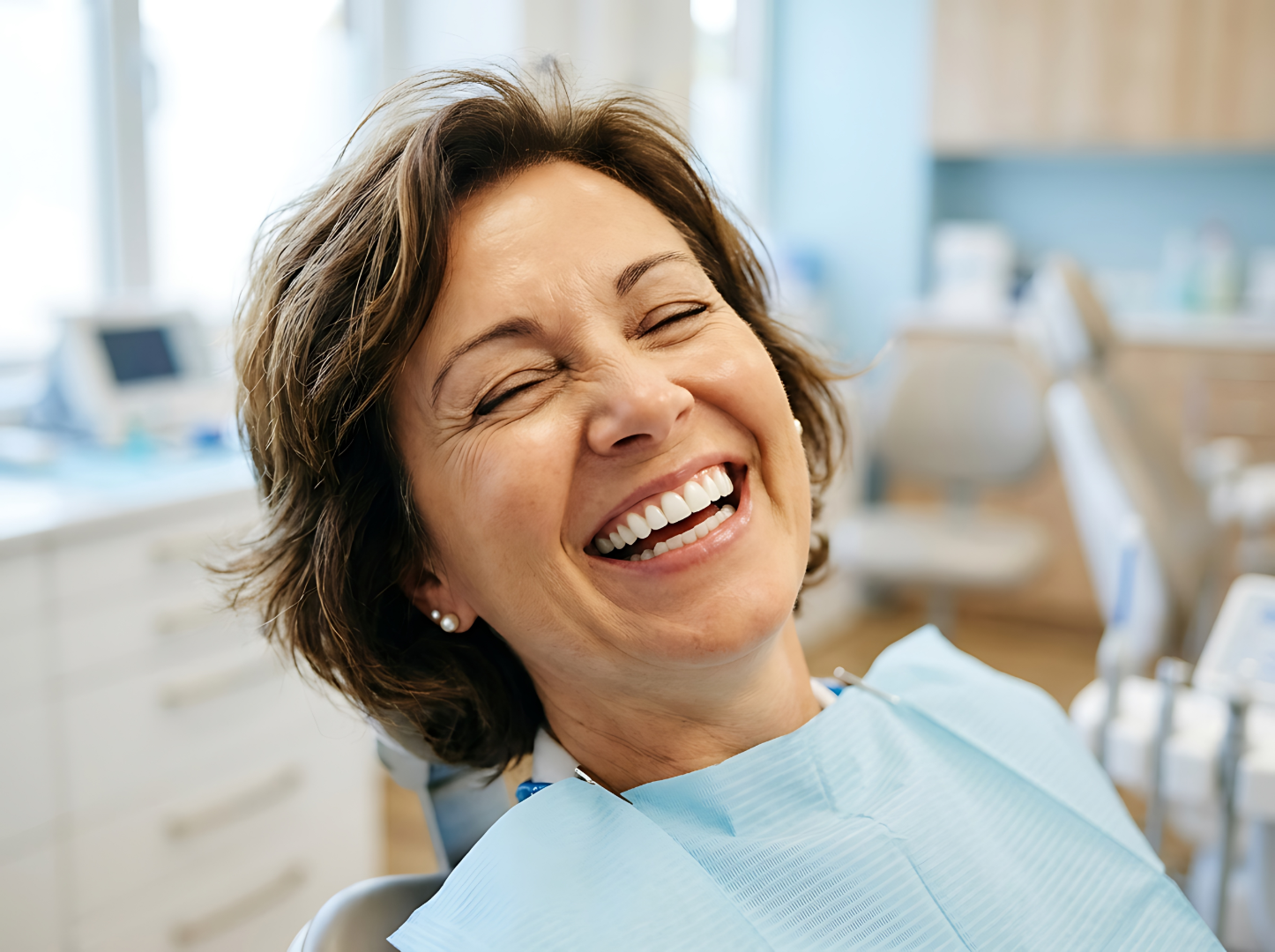 Happy woman with pearl earrings laughing in a dental clinic wearing a blue dental bib.