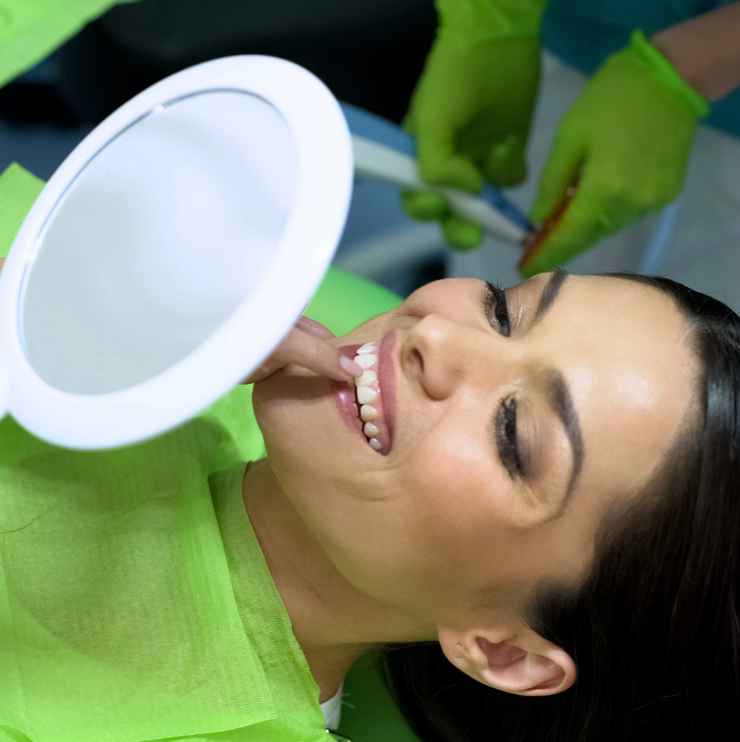 Woman wearing a green dental bib smiles looking at her teeth in a handheld mirror during dental care.