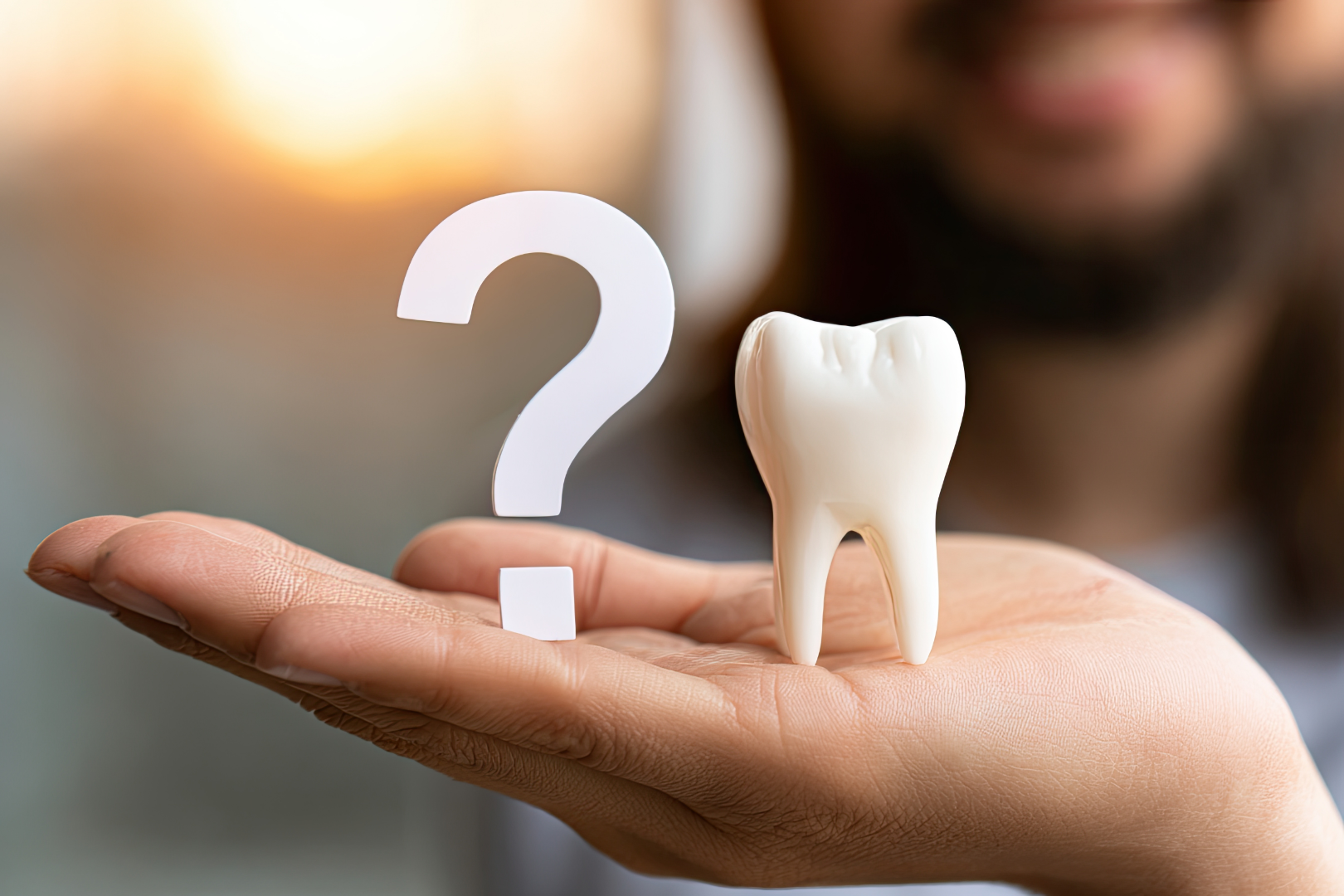 Hand holding a model tooth and a white question mark symbol with blurred face background.