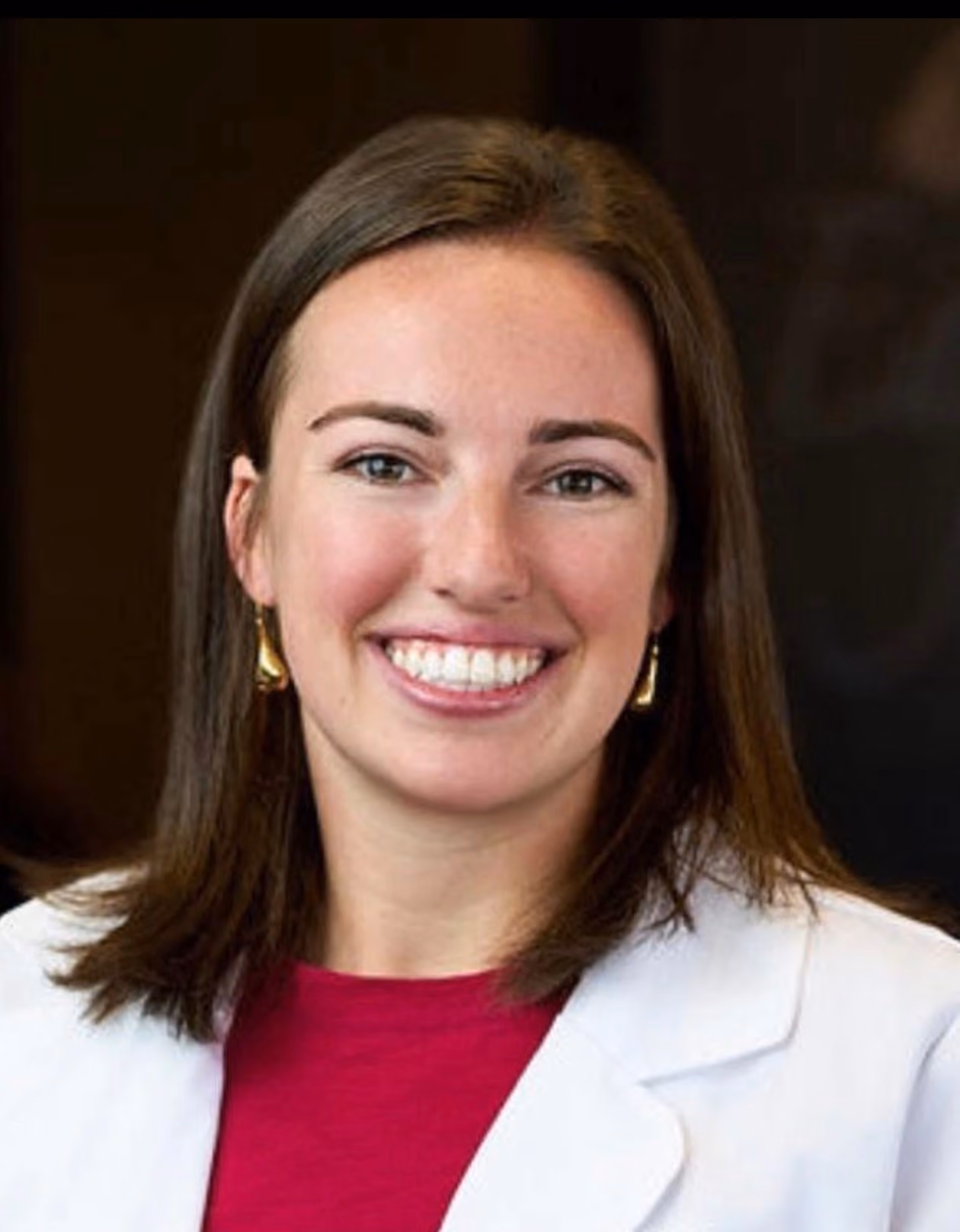 Smiling woman with shoulder-length brown hair, wearing gold earrings, a red top, and a white coat.