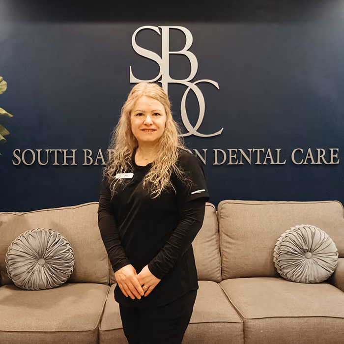Smiling woman in black scrubs standing in front of a couch and dental care sign on navy wall.