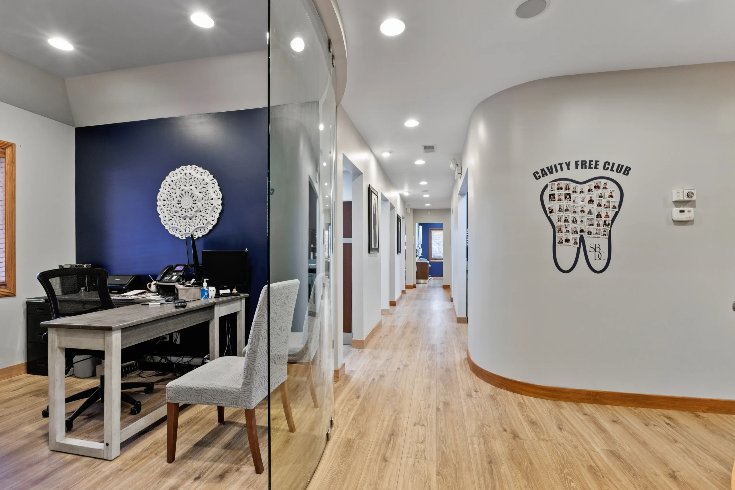 Modern dental office hallway with wood flooring, reception desk, and 'Cavity Free Club' tooth photo wall.