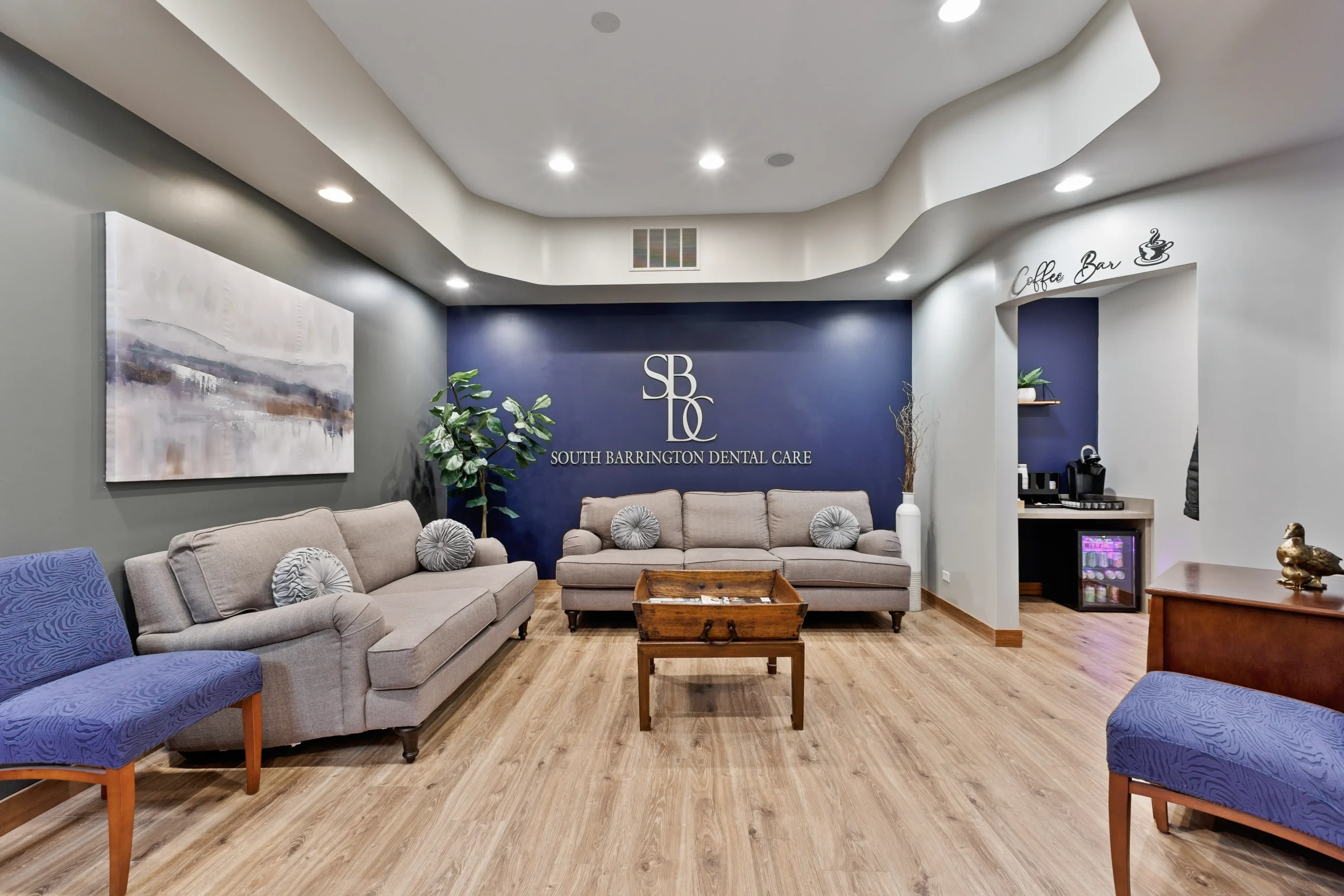 Modern dental clinic waiting room with sofas, blue chairs, wooden table, and coffee bar corner.