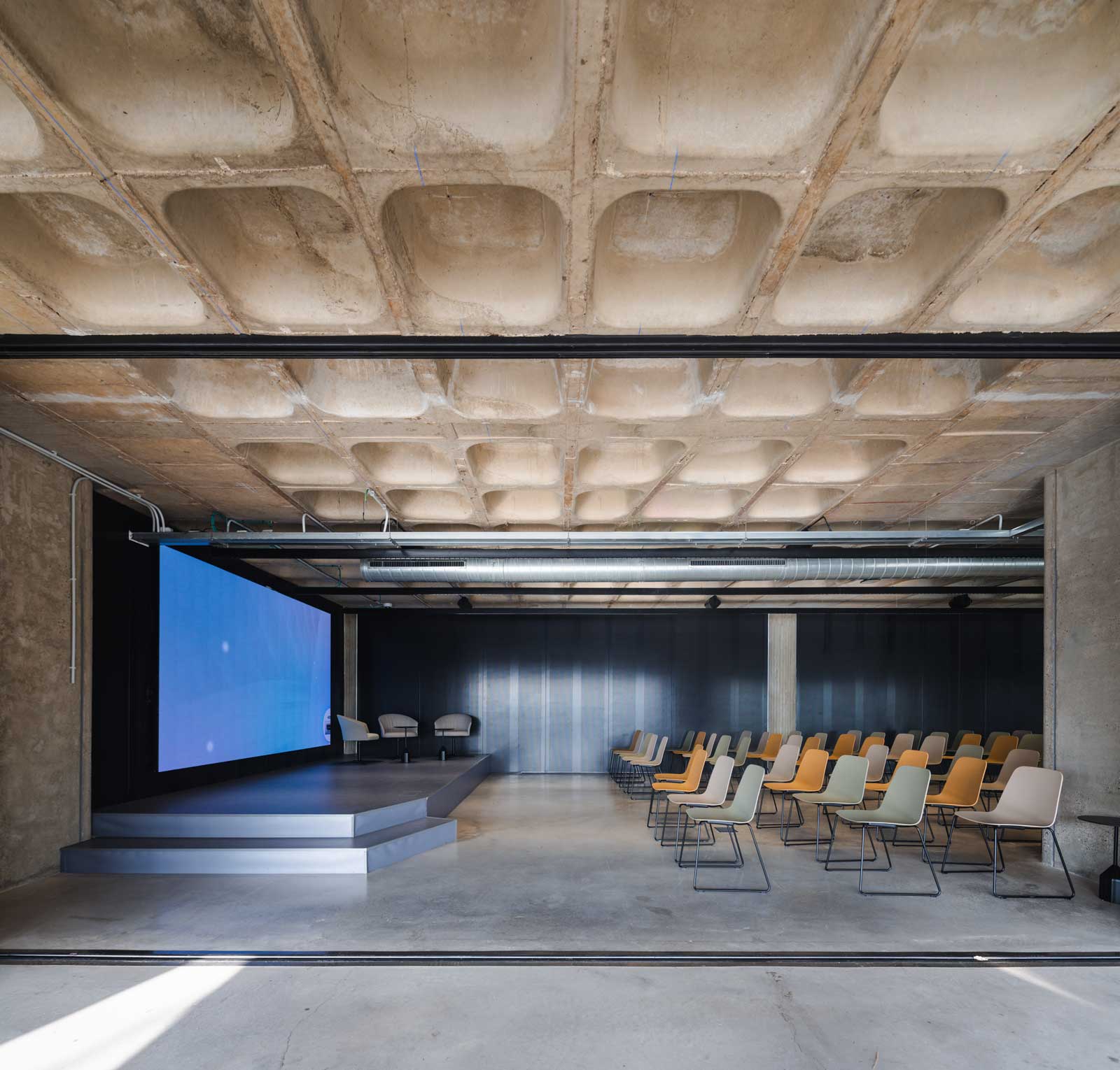 Sala de conferencias moderna con sillas en tonos neutros y escenario con tres sillones y pantalla azul.