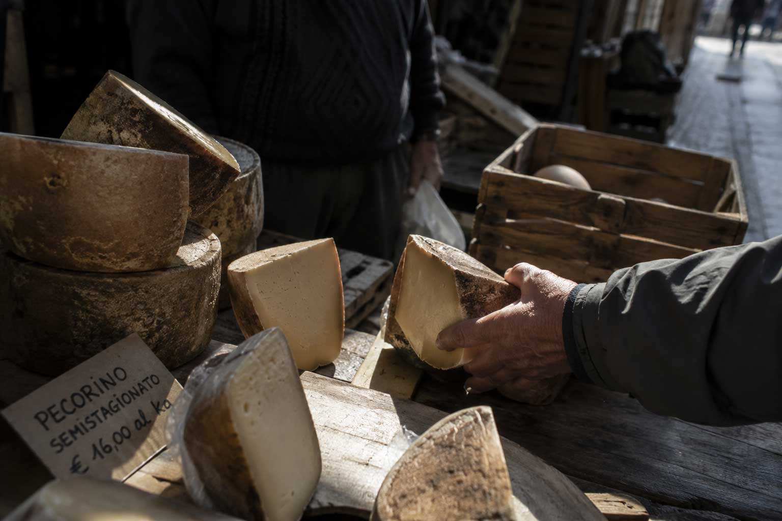 Hand holding a wedge of aged Pecorino cheese at an outdoor market stall with wooden crates.