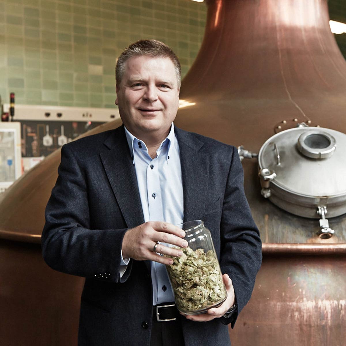 Vorstand der PARK und Bellheimer AG Roald Pauli hält ein Glas mit Hopfen vor einem großen Kupferbraukessel in der Brauerei von PARK und Bellheimer.