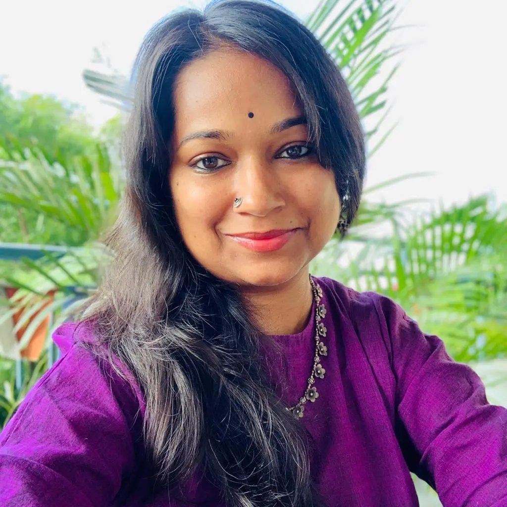 Image of Vasvi wearing a purple top and silver jewelry smiling outdoors near green plants.