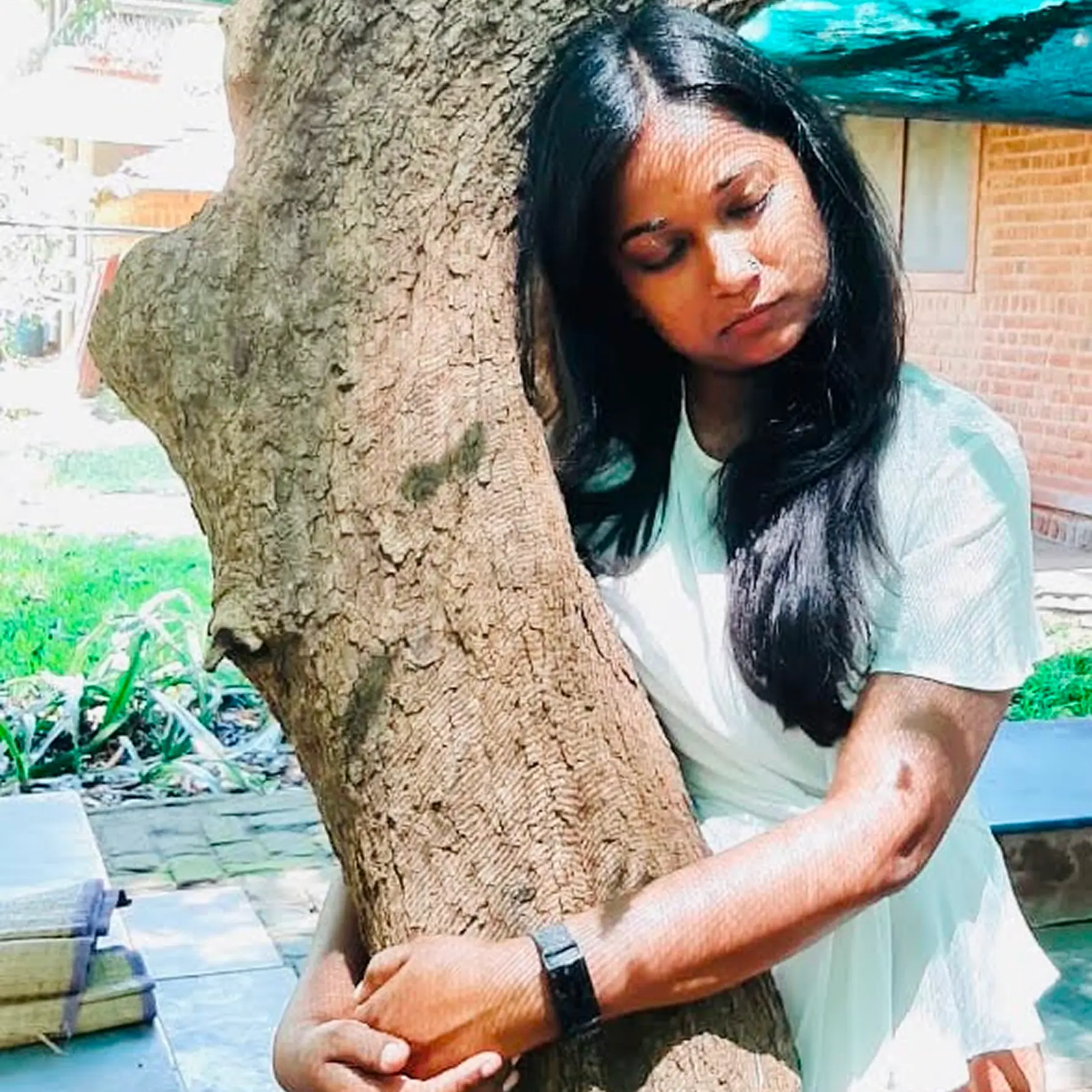 Vasvi in white hugging a tree trunk outdoors with her eyes closed in a peaceful setting.