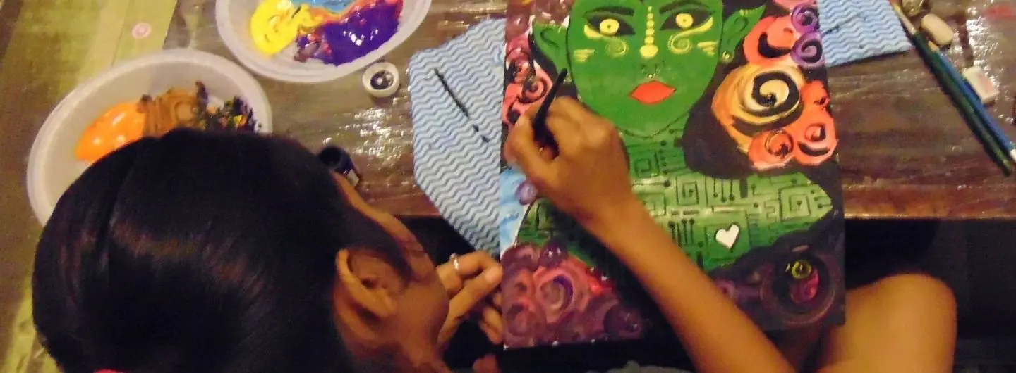 Person painting a colorful green face on canvas with swirling patterns and yellow eyes.